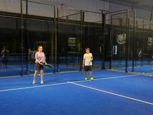 Uczennica i uczeń ćwiczą odbicia na korcie padlowym, stojąc po przeciwnych stronach siatki.