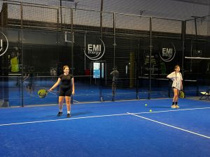 Dwie uczennice stoją na korcie padlowym z rakietami, czekając na rozpoczęcie gry lub wskazówki instruktora.