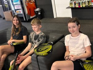 Trójka uczniów siedzi na kanapie w klubie sportowym, trzymając rakiety do padla i odpoczywając po zajęciach.