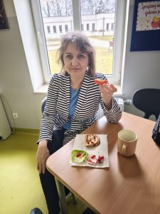 Nauczycielka w szlafroku siedzi przy stoliku i je przekąskę podczas szkolnych obchodów Dnia w Piżamie.