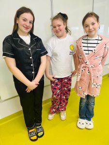 Trzy uczennice ubrane w piżamy stoją w klasie i uśmiechają się podczas obchodów Dnia w Piżamie w szkole.