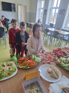 Dzieci w piżamach stoją przy stole z przygotowanymi zdrowymi przekąskami w szkolnej stołówce podczas Dnia w Piżamie.
