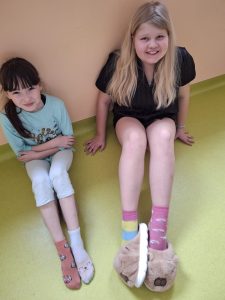 Dwie uczennice siedzą pod ścianą i prezentują różne, kolorowe skarpetki jako symbol solidarności ze Światowym Dniem Downa.