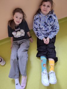 Dwie uczennice siedzą pod ścianą i pokazują kolorowe, niepasujące skarpetki oraz kapcie, solidaryzując się ze Światowym Dniem Downa.