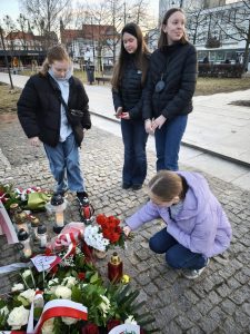 Uczennica Samorządu Uczniowskiego składa kwiaty przy pomniku Ofiar Wyklętych, a pozostałe uczennice stoją obok podczas uroczystości 1 marca.