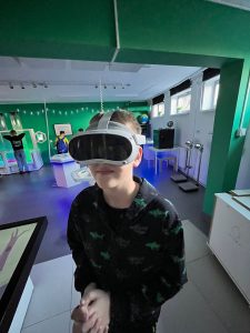 Uczeń stoi w Laboratorium Wyobraźni i korzysta z gogli wirtualnej rzeczywistości, w tle widoczne interaktywne stanowiska edukacyjne.