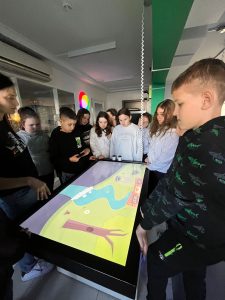 Grupa uczniów wraz z edukatorem korzysta z interaktywnego stołu edukacyjnego przedstawiającego animowaną planszę.