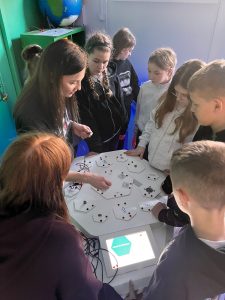 Uczniowie pod kierunkiem edukatora budują obwód elektryczny na interaktywnym stanowisku.