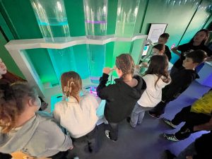 Grupa uczniów eksperymentuje przy stanowisku z przezroczystymi cylindrami wypełnionymi kolorową cieczą.