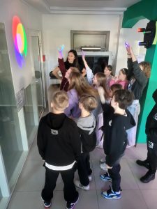 Grupa uczniów wraz z edukatorem obserwuje doświadczenie z mieszaniem kolorów światła na ścianie.