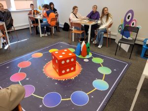 Uczniowie siedzą przy stolikach i uczestniczą w edukacyjnej grze o tematyce kosmicznej. Na pierwszym planie widoczna jest plansza edukacyjnej gry kosmicznej z dużą kostką i modelami rakiet na środku.