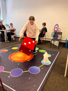 Uczennica uczestniczy w edukacyjnej grze o tematyce kosmicznej, rzucając dużą kostką na planszy.