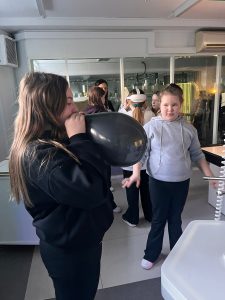 Uczennica nadmuchuje balon podczas eksperymentu z powietrzem, inni uczniowie obserwują doświadczenie.