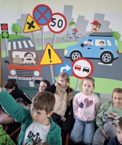 Grupa dzieci siedzi przed planszą edukacyjną ze znakami drogowymi podczas zajęć w WORD.