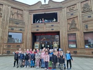 Grupa dzieci stoi przed wejściem do Teatru Baj Pomorski podczas wycieczki na spektakl „Szewczyk Dratewka”.