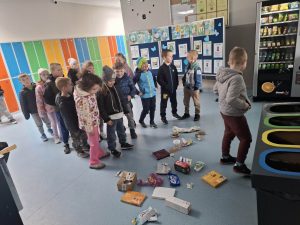 Dzieci ustawione w kolejce segregują odpady do odpowiednich pojemników w szkolnym korytarzu.