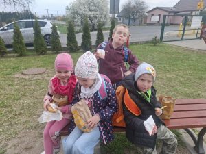 Grupa dzieci siedzi na ławce na świeżym powietrzu i je przekąski po powrocie z wycieczki.