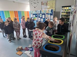 Grupa dzieci uczestniczy w ćwiczeniu segregacji śmieci, wrzucając odpady do oznaczonych koszy.