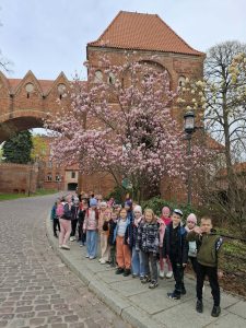 Dzieci ustawione wzdłuż chodnika pozują do zdjęcia przy kwitnącym drzewie i zabytkowym budynku.