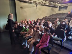 Dzieci siedzą na widowni sali teatralnej i słuchają opiekuna wyjazdu przed spektaklem.