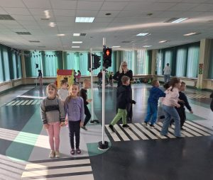 Dzieci przechodzą przez oznaczone przejście dla pieszych w sali edukacyjnej, ucząc się zasad ruchu drogowego.