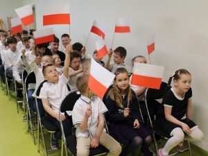 Uczniowie siedzący na krzesłach trzymają małe biało-czerwone flagi w czasie apelu.