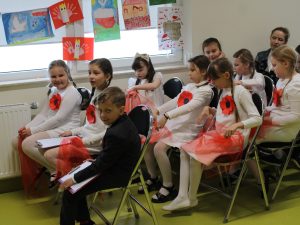 Grupa dzieci siedzi w rzędach, część z nich przygotowuje elementy stroju do występu.