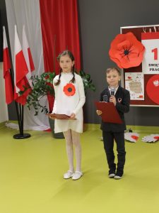 Dziewczynka i chłopiec stoją na środku sali; chłopiec mówi do mikrofonu, dziewczynka trzyma kartkę.