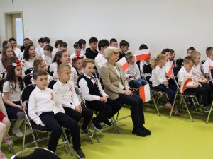 Publiczność szkolna siedzi na krzesłach, uczniowie trzymają biało-czerwone flagi. Wśród uczniów siedzi pani dyrektor.