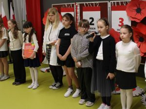 Grupa uczniów stoi na scenie; jedna dziewczynka mówi do mikrofonu, inni czekają na swoją kolej.