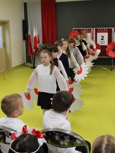 Dzieci wchodzą w szeregu, trzymając dekoracyjne elementy przypominające biało-czerwone kwiaty.