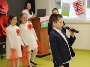 Dziewczynki ustawione w rzędzie trzymają czerwone wstążki, przygotowane do występu tanecznego. Na pierwszym planie widoczny jest chłopiec stojący bokiem i mówiący do mikrofonu.