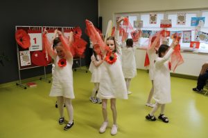 Grupa dziewczynek wykonuje choreografię, machając czerwonymi wstążkami w sali udekorowanej na święta majowe.
