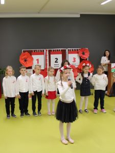 Najmłodsze dzieci stoją na scenie; jedna dziewczynka śpiewa lub recytuje do mikrofonu.