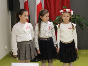 Trzy dziewczynki w odświętnych strojach stoją przed biało-czerwonymi flagami podczas szkolnego apelu.