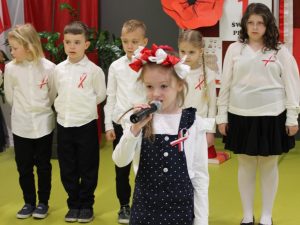 Dziewczynka mówi do mikrofonu, za nią stoi grupa uczniów w galowych strojach z biało-czerwonymi przypinkami.