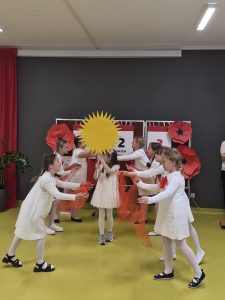Dzieci tworzą układ taneczny wokół koleżanki trzymającej dużą dekorację w kształcie słońca, używając czerwonych chust.