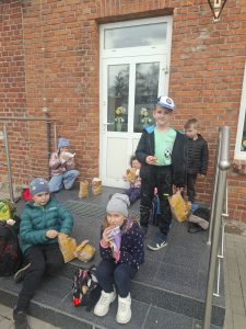 Dzieci z oddziału przedszkolnego i klasy pierwszej siedzą na schodach przed budynkiem szkoły i jedzą posiłek po powrocie z wycieczki.