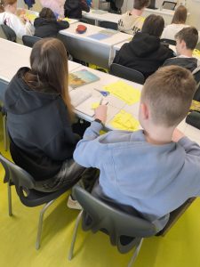 Uczniowie siedzą tyłem w klasie i pracują nad wycinaniem żonkili z żółtego papieru, przygotowując elementy do wspólnej pracy plastycznej.