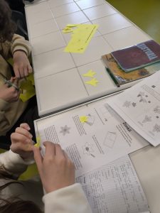 Uczniowie siedzą przy ławkach i wycinają z żółtego papieru kształty żonkili, korzystając z nożyczek i instrukcji leżących na stole.