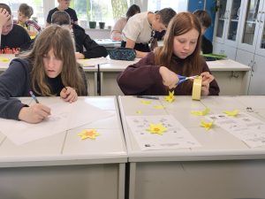 Dwie uczennice przy ławce wycinają i przygotowują papierowe żonkile, wokół nich leżą materiały plastyczne i gotowe elementy kwiatów.