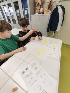 Dwóch uczniów siedzi przy stole i pracuje nad papierowymi żonkilami, korzystając z instrukcji i przyborów szkolnych.