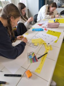 Trzy uczennice siedzą przy stole i przygotowują papierowe żonkile; na blacie widoczne są nożyczki, klej i kolorowe pisaki.