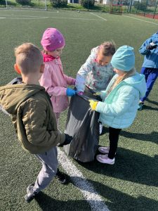 Uczniowie w rękawicach stoją na boisku i wspólnie zbierają śmieci do czarnego worka podczas szkolnej akcji sprzątania z okazji Dnia Ziemi.
