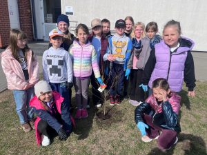 Grupa dzieci stoi wokół świeżo posadzonego drzewka i pozuje do zdjęcia przed budynkiem szkoły.