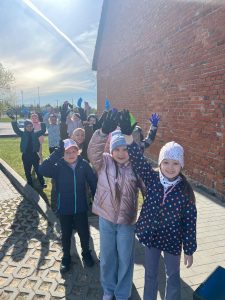 Grupa młodszych dzieci idzie chodnikiem i podnosi ręce w górę, pokazując rękawiczki używane podczas prac ogrodowych.