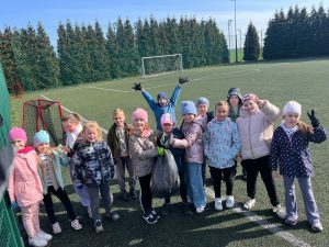 Grupa uczniów na boisku szkolnym prezentuje zebrany worek odpadów; dzieci uśmiechają się i pozują po zakończonym sprzątaniu terenu szkoły.