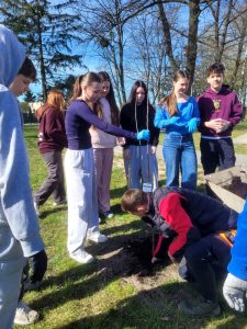 Uczniowie szkoły sadzą drzewko na terenie zielonym przy budynku; jedna osoba umieszcza sadzonkę w dołku, pozostali obserwują i pomagają.