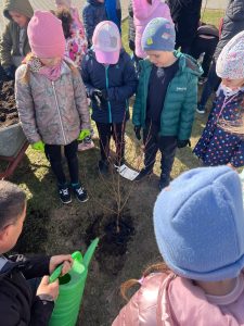 Dzieci stoją wokół świeżo posadzonego drzewka, a dorosły podlewa je zieloną konewką.
