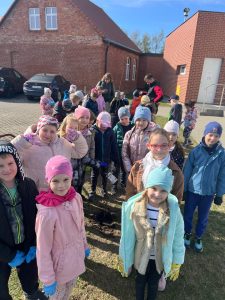 Duża grupa dzieci stoi wokół nowo posadzonego drzewka na terenie szkoły i pozuje do zdjęcia.